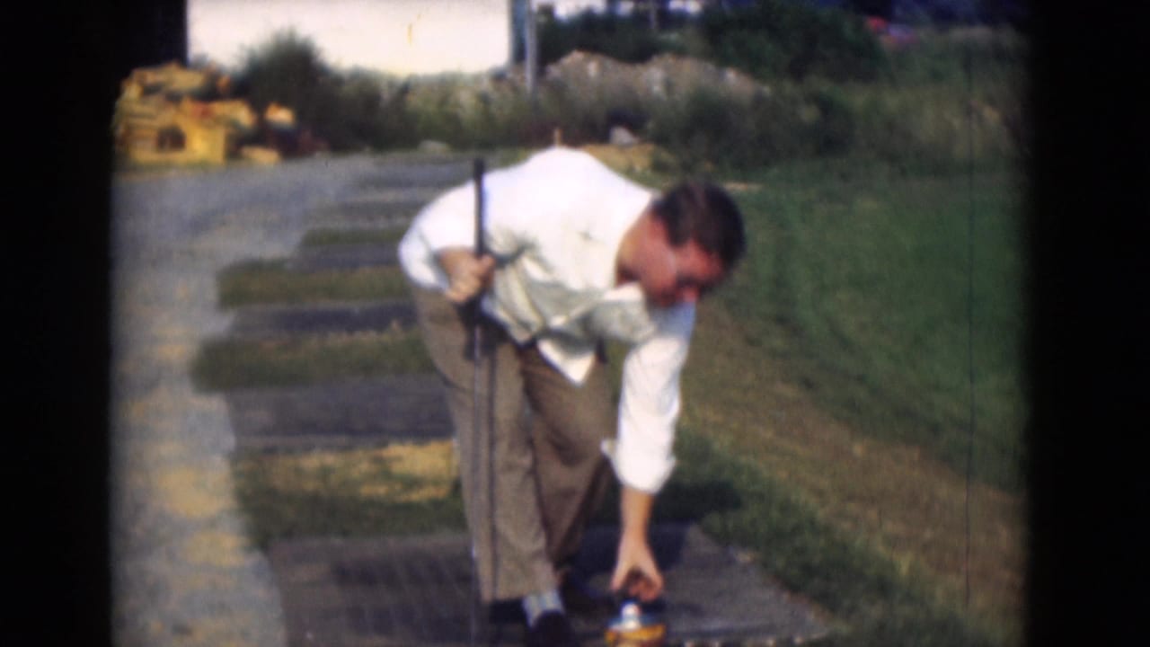 Man in a white shirt bends toward a golf ball while holding a club on grass near a path.