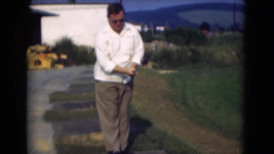A man in white shirt and brown trousers holds his golf club on a grassy course area in the Middletown, Pennsylvania 1975 footage.