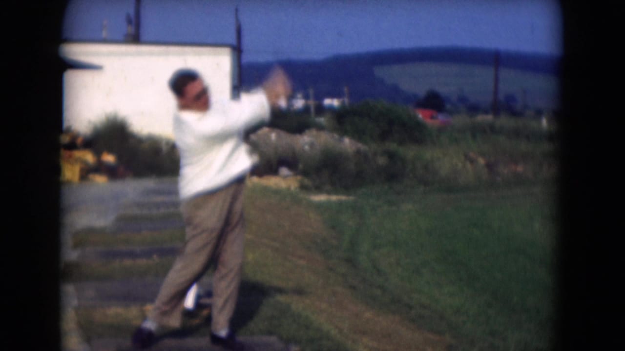 The 1975 home-movie frame shows the man mid-swing, turning his body as the golf club rises against a bright outdoor backdrop.