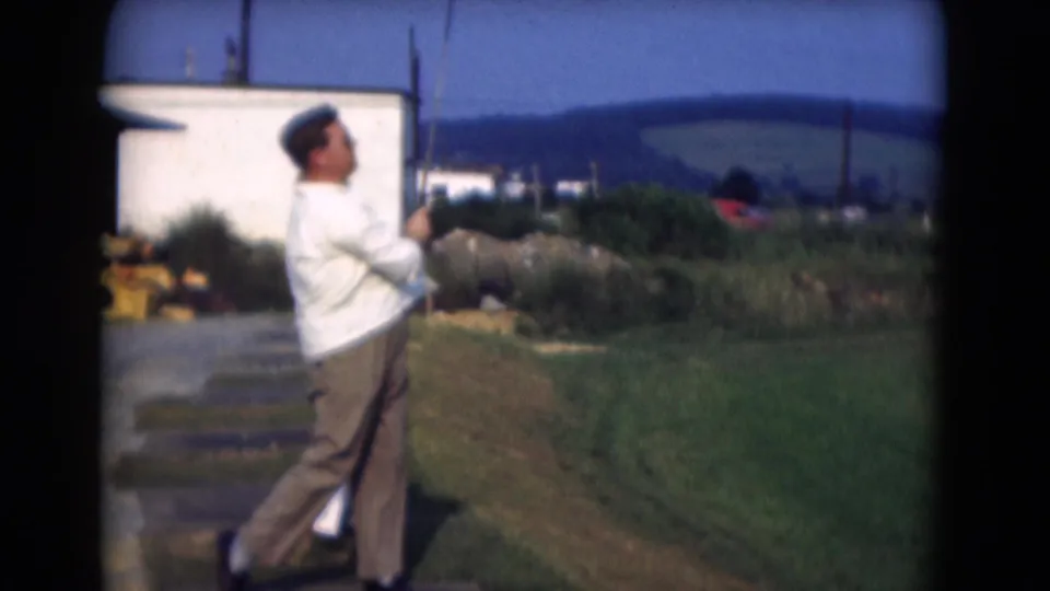 In the supported Middletown 1975 setting, the man continues his golf follow-through beside grass, a paved path, and a white structure.