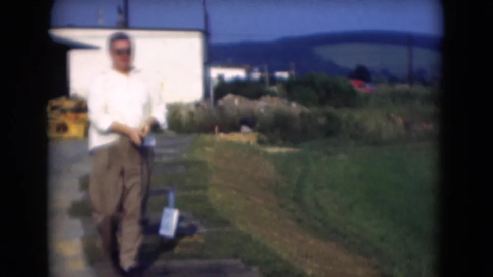 The man lowers the golf club at the end of the short 8mm sequence, standing in bright sun with grass and hills behind him.