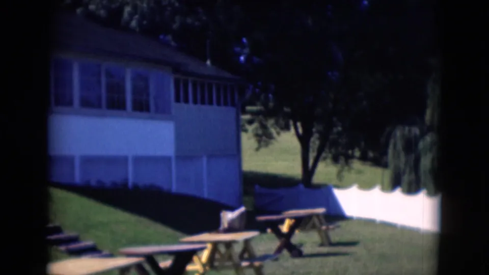 1975 Middletown, Pennsylvania home-movie frame showing a white house, grassy yard, picnic tables, and trees in bright sun.