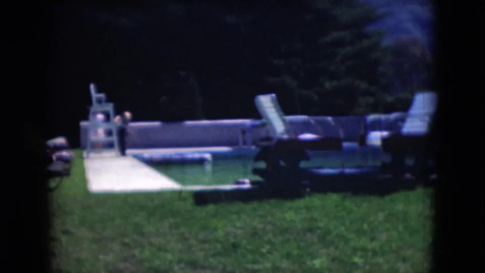 Middletown, Pennsylvania 1975 home-movie frame showing a backyard pool, lounge chairs, white fence, lawn, and distant person.