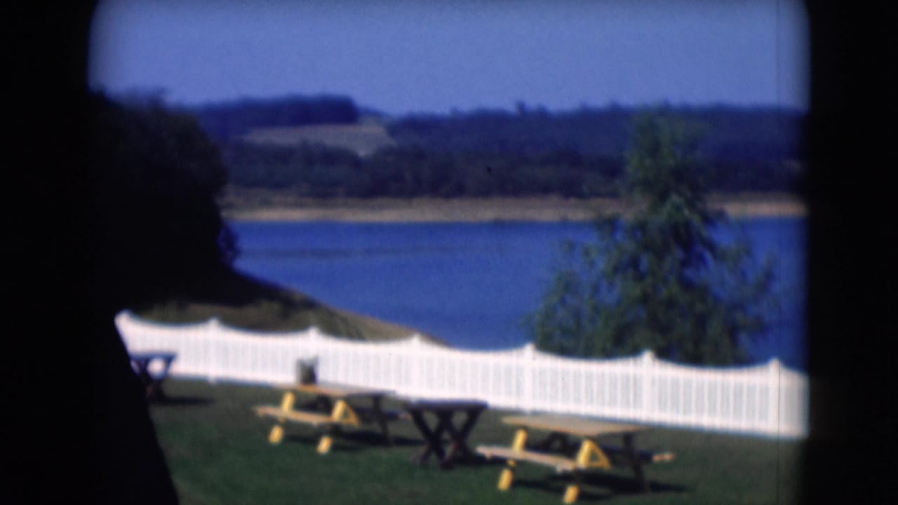 1975 Middletown, Pennsylvania 8mm frame showing a lakeside yard with picnic tables, a white fence, blue water, and wooded hills.