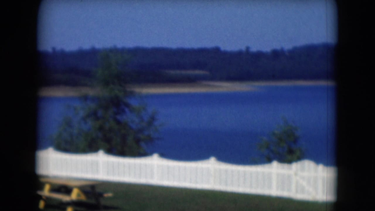 1975 Middletown, Pennsylvania lakeside view with a white fence, shrubs, blue water, and distant wooded shoreline.