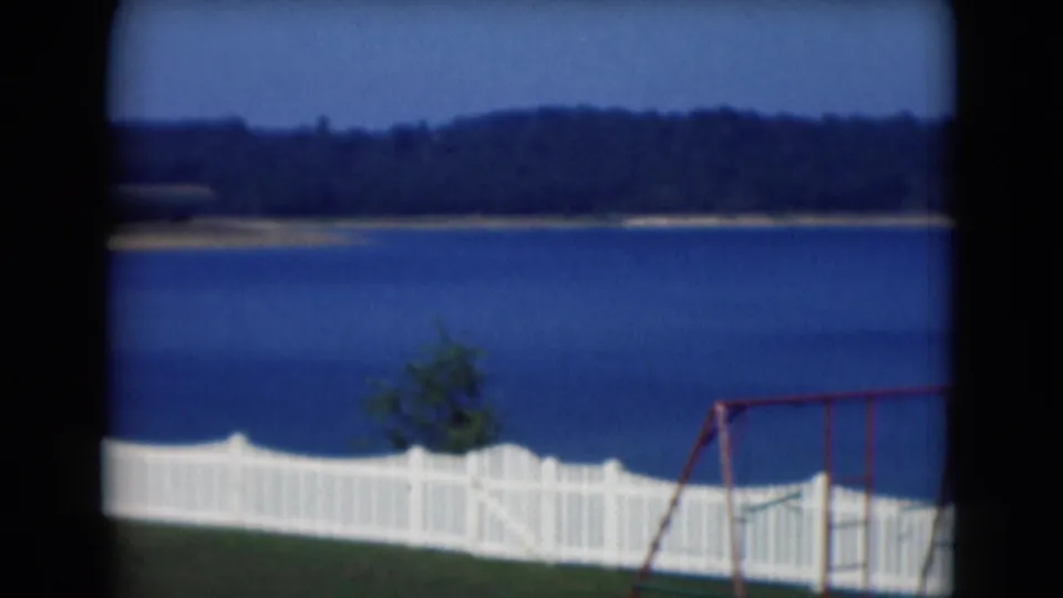 1975 Middletown, Pennsylvania home-movie frame with a white lakeside fence and red swing set near calm blue water.