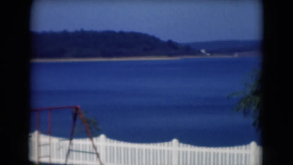 1975 8mm view of a blue lake near Middletown, Pennsylvania, seen past a white fence and red swing set.