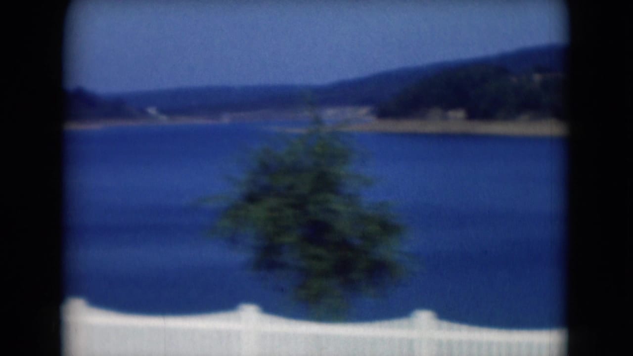 1975 8mm lakeside view near Middletown, Pennsylvania, with a white fence, blue water, distant hills, and foreground greenery.