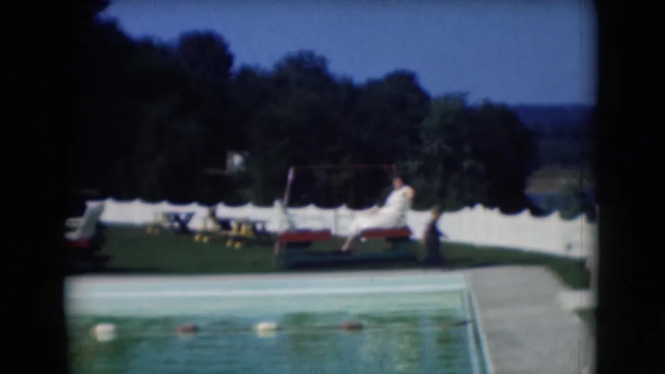 1975 8mm poolside scene near Middletown, Pennsylvania, showing a person reclining near lounge chairs, a white fence, and trees.