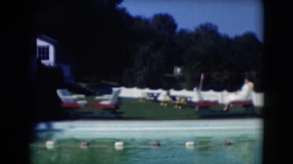 1975 8mm view of a poolside lawn near Middletown, Pennsylvania, with lounge chairs, trees, a white fence, and a distant person.