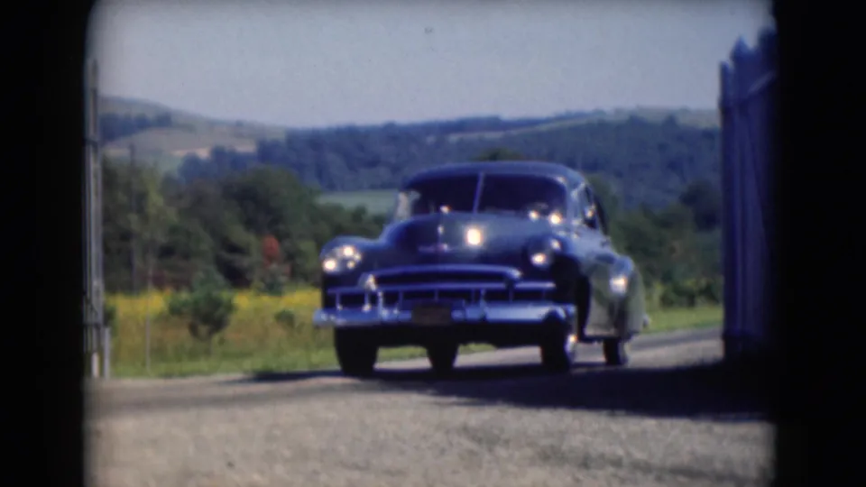 A dark mid-century sedan drives closer along a rural Middletown road, framed by a white gate and distant hills.
