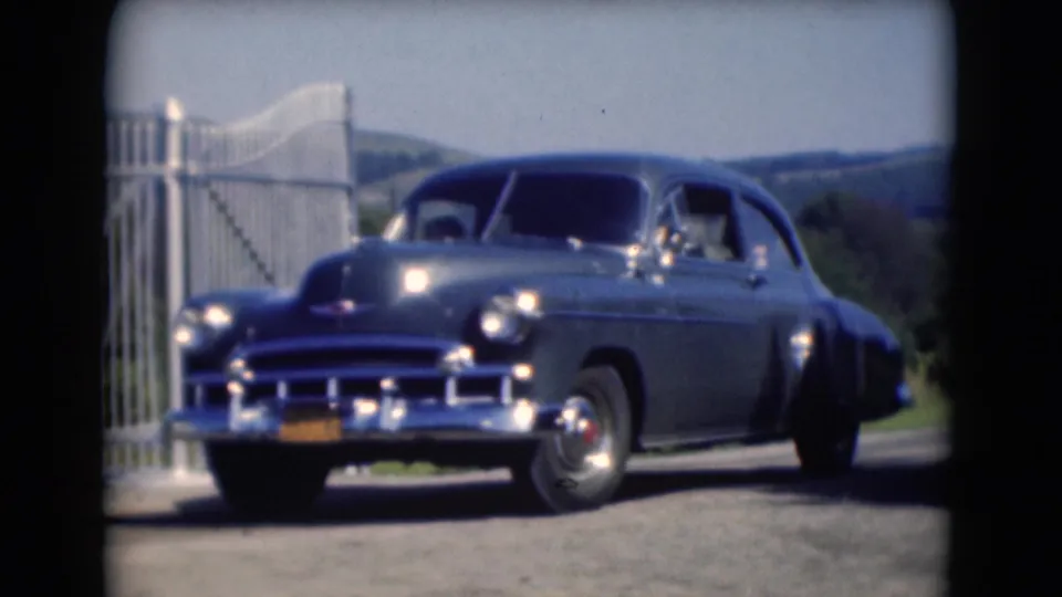 A dark vintage Chevrolet-style sedan passes the camera near a white gate in rural Middletown, with a person faintly visible inside.
