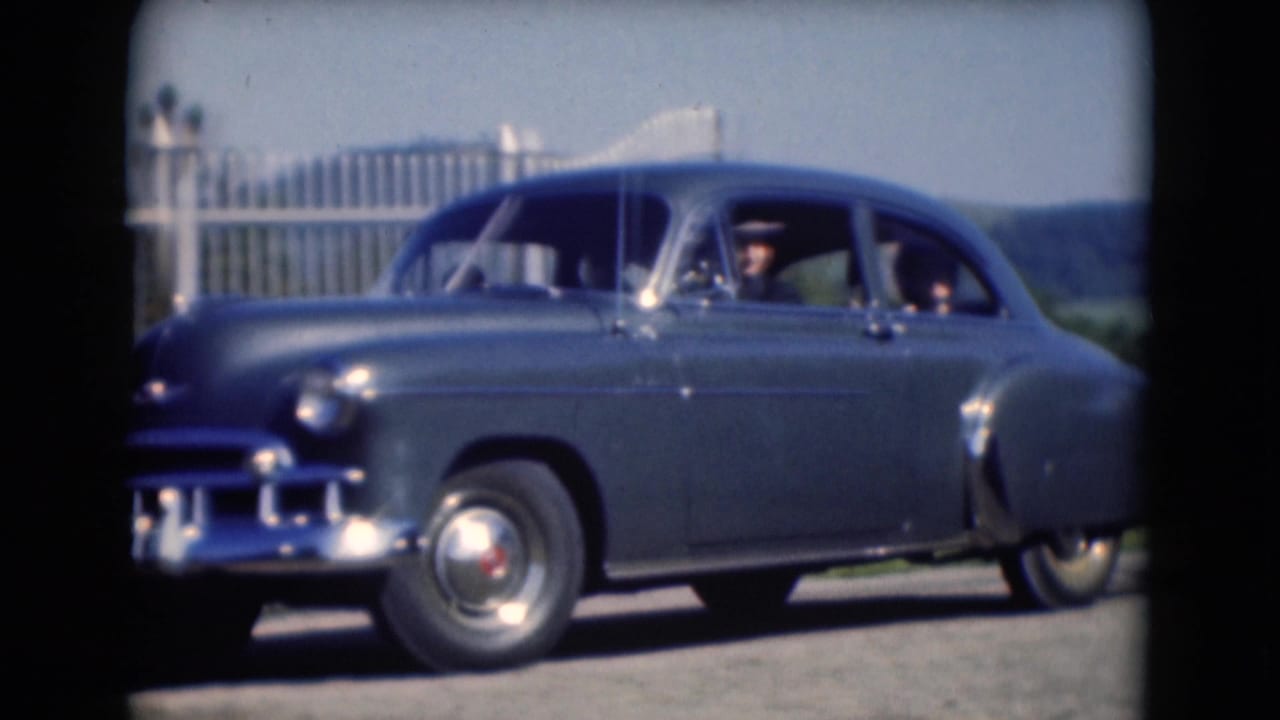 Blue vintage sedan passing a roadside camera with driver and passenger visible.