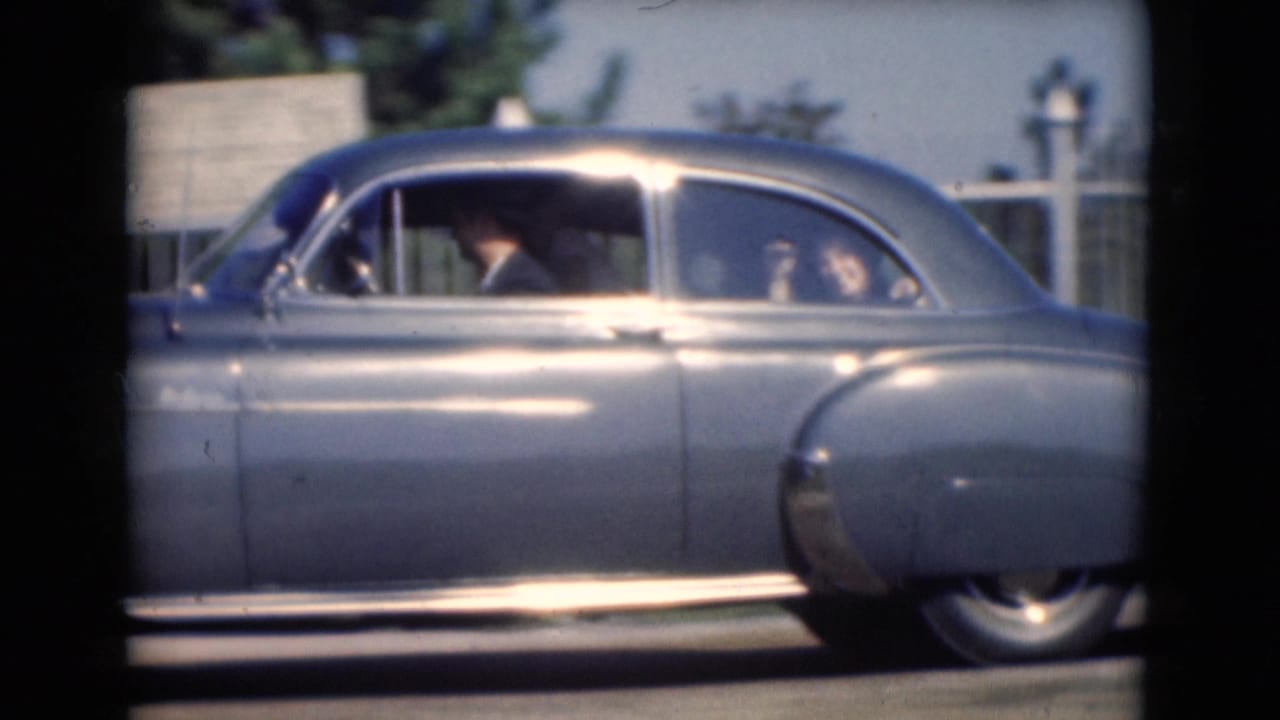 A blue mid-century sedan fills the frame in 1975 Middletown footage, with a driver and passenger seen through the windows.
