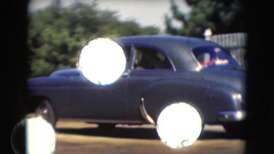 The sedan continues past the camera, seen from the rear side with passengers faintly visible and bright 8mm flare over the car body.