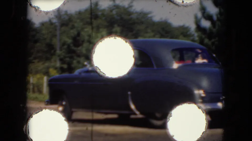 The blue sedan exits frame right in the 1975 home-movie scene, with rear passengers faintly visible behind strong circular flare artifacts.