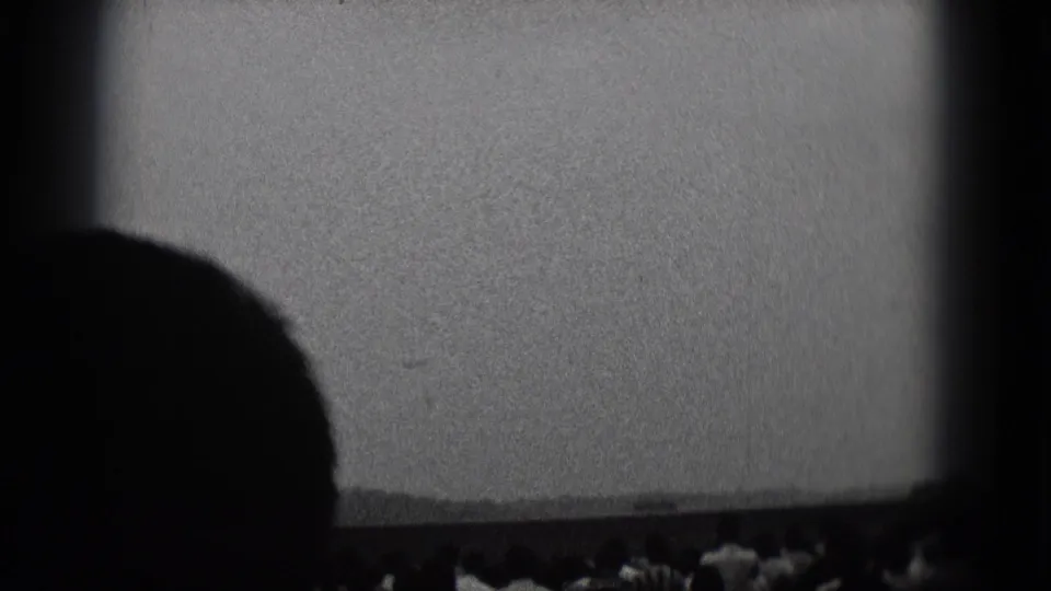 1975 black-and-white home-movie frame from a theater, with a large seated silhouette below the blank screen.