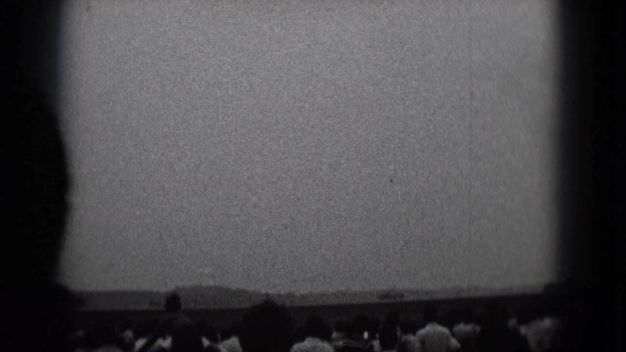 Crowd silhouettes line the lower frame beneath a bright sky and distant low hills.