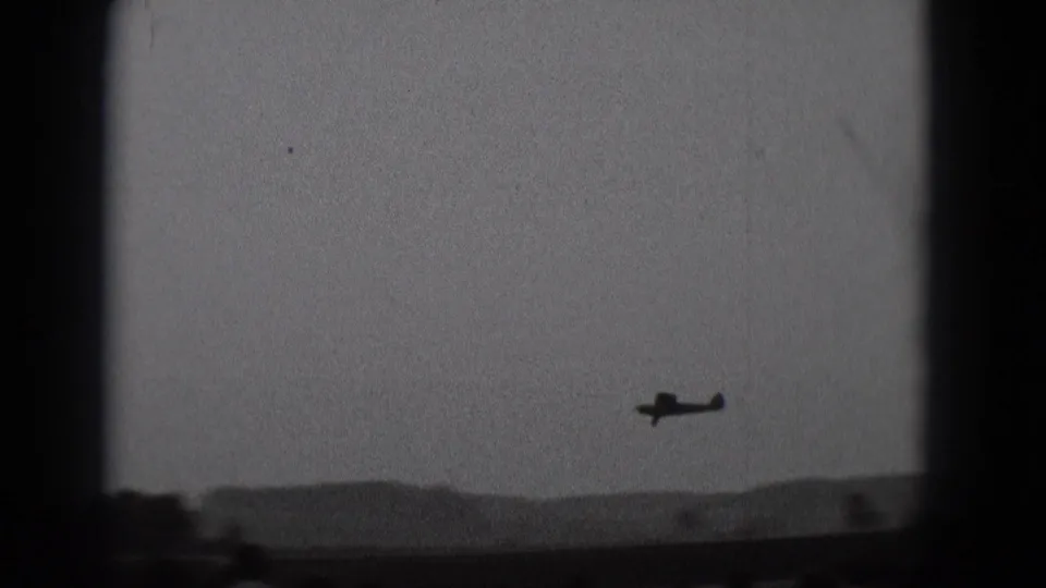 1975 Middletown, Pennsylvania 8mm frame showing a small airplane in silhouette crossing a hazy sky above a low horizon.