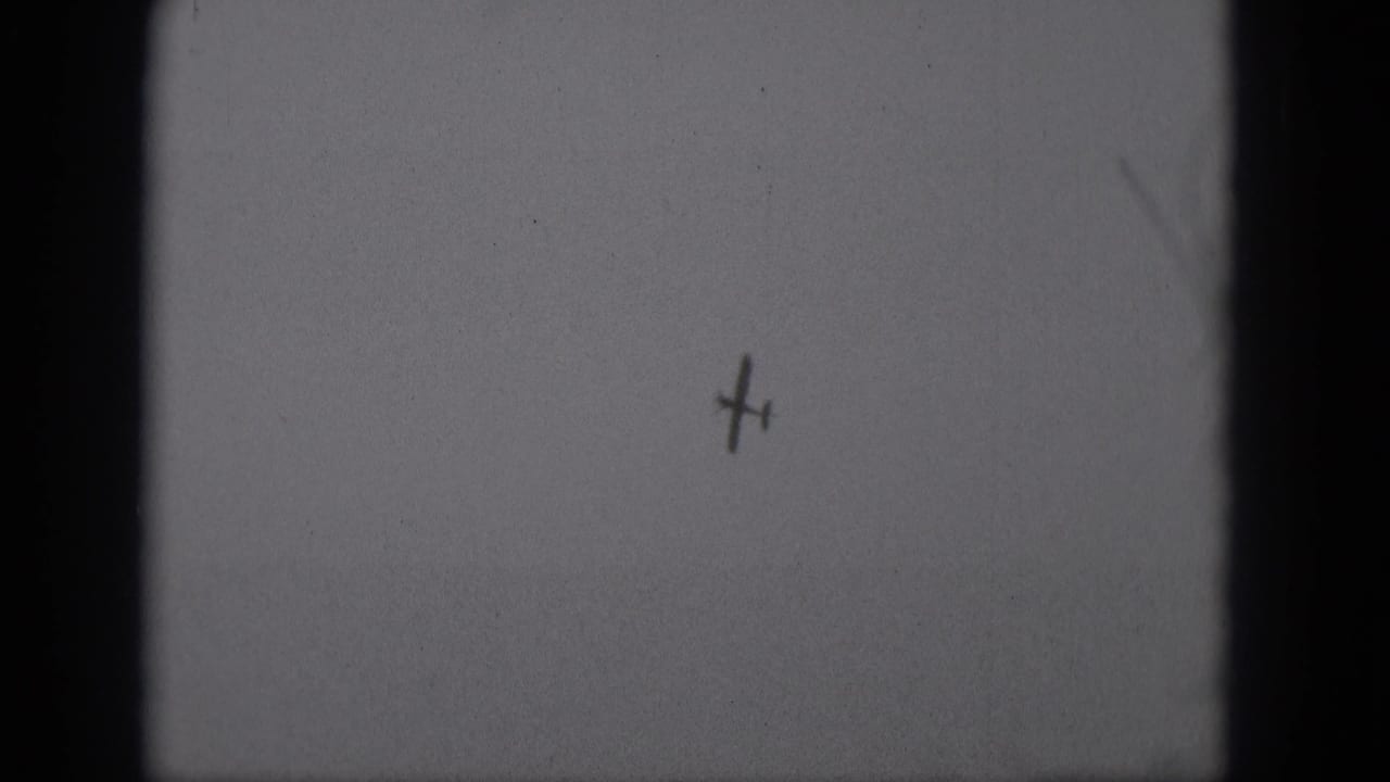 Small dark aircraft silhouette against a pale overcast sky with heavy film grain and gate shadows.