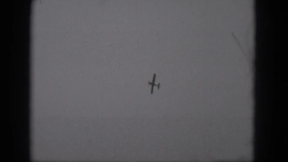 1975 8mm home-movie frame from Middletown, Pennsylvania, showing a tiny aircraft silhouette high in a pale sky.
