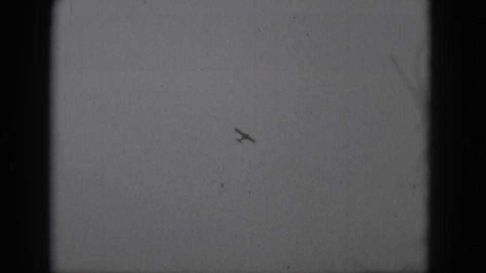 Monochrome 8mm frame showing the small airplane lower in the sky, with visible grain, dust and dark gate edges.