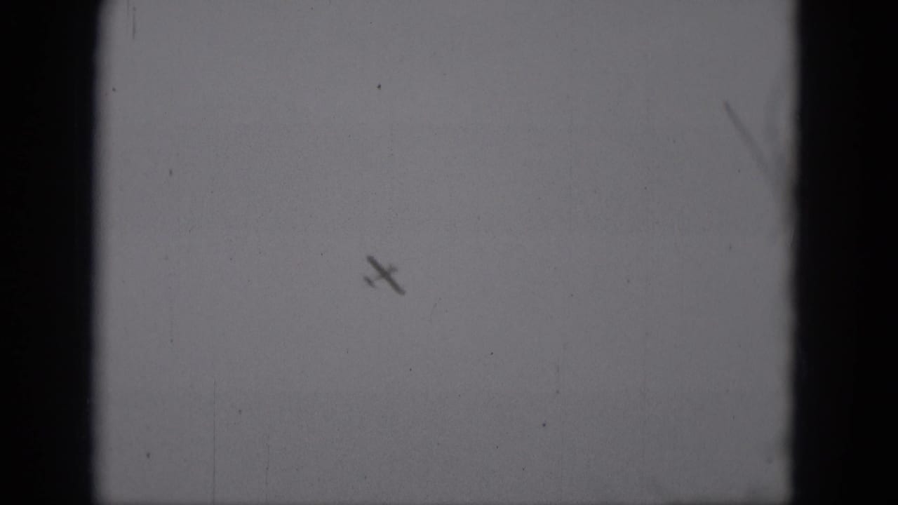 Small airplane silhouette banking against a pale sky with visible film grain.
