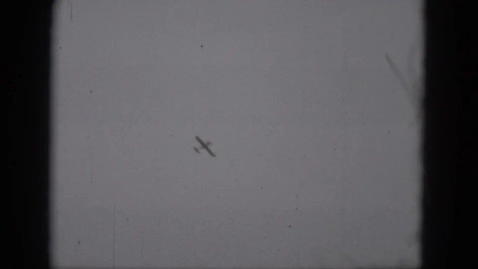 1975 8mm home-movie frame from Middletown, Pennsylvania, showing a small airplane silhouette banking high against a pale sky.