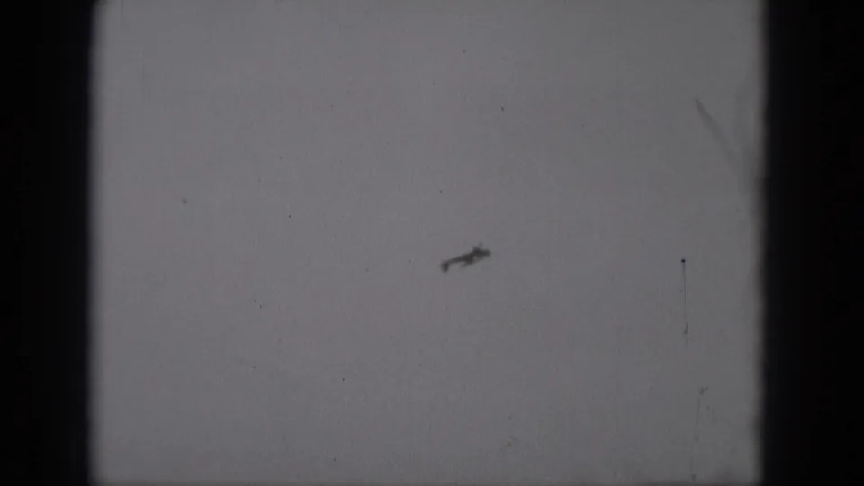 The small plane appears lower in the frame, reduced to a dark silhouette against grainy 8mm sky footage from Middletown in 1975.