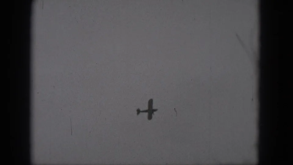 A small airplane silhouette appears to rotate or bank steeply against a pale gray sky in the closing 1975 home-movie frame.
