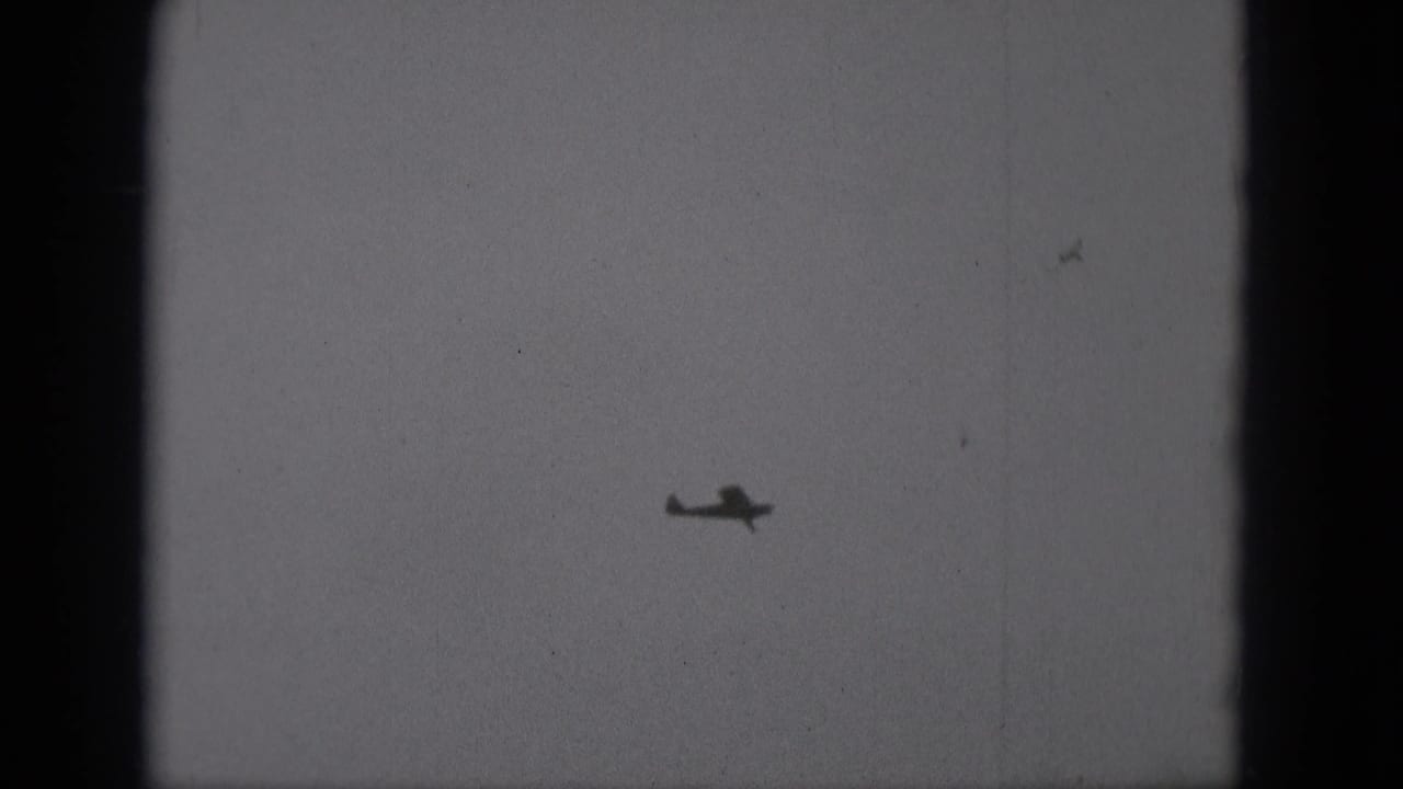 Small airplane silhouette in a pale overcast sky with dark film-frame edges.