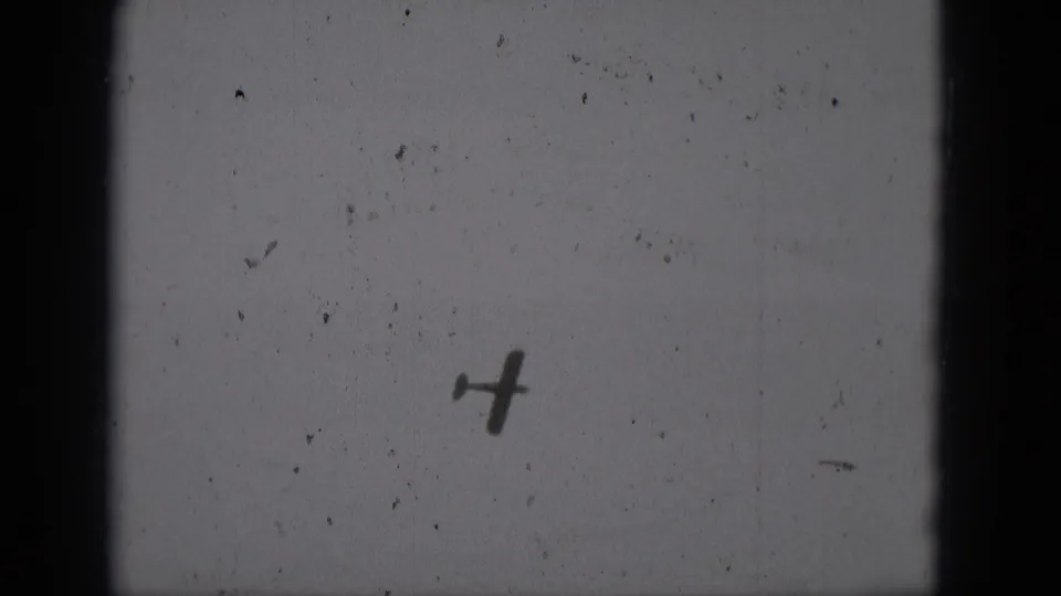 A small propeller plane silhouette sits low in the frame against a gray sky, with dust and gate edges visible in the 8mm scan.