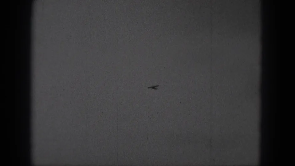 The airplane appears smaller and farther away against the overcast sky, with the 8mm frame edges and grain still visible.