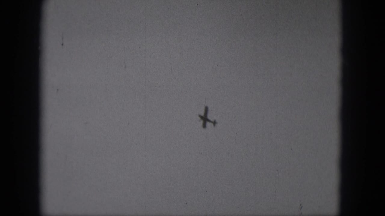 A distant small plane appears as a dark silhouette in a pale sky, from 1975 Middletown, Pennsylvania 8mm footage.