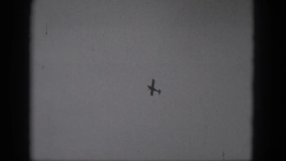 A distant small plane appears as a dark silhouette in a pale sky, from 1975 Middletown, Pennsylvania 8mm footage.