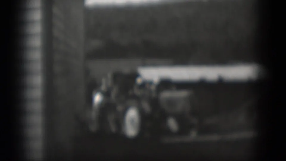 1975 Middletown home-movie frame showing people beside a parked tractor-like vehicle near a light-sided rural building.