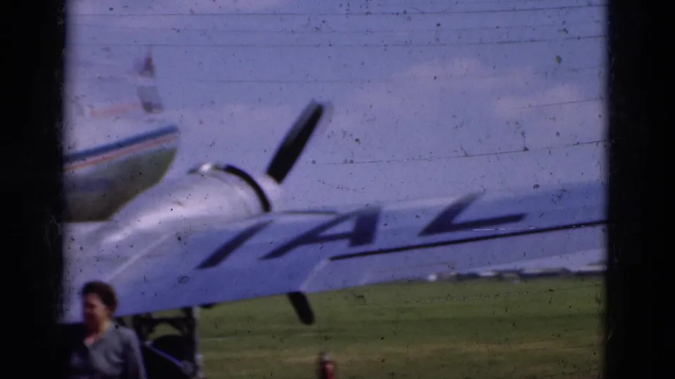 1975 Portugal home-movie frame showing a propeller aircraft on grass with IAL lettering and an adult figure near the wing.