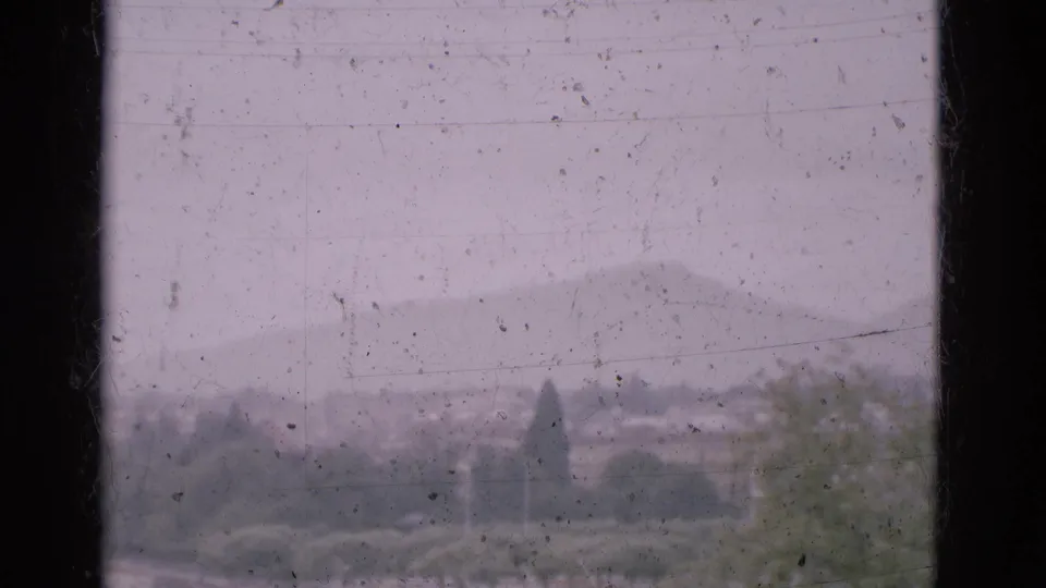 1975 Portugal home-movie frame with a soft, hazy view of trees, distant buildings, and mountains under a pale sky.