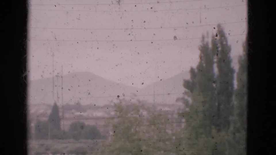 The camera drifts across a dusty window view as tall trees and a faint mountain ridge appear in 1975 Portugal footage.