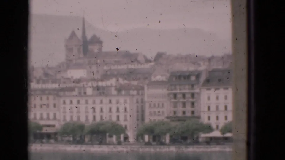 1975 Portugal 8mm view of waterfront buildings, a tree line, and distant twin spires seen through dusty window glass.
