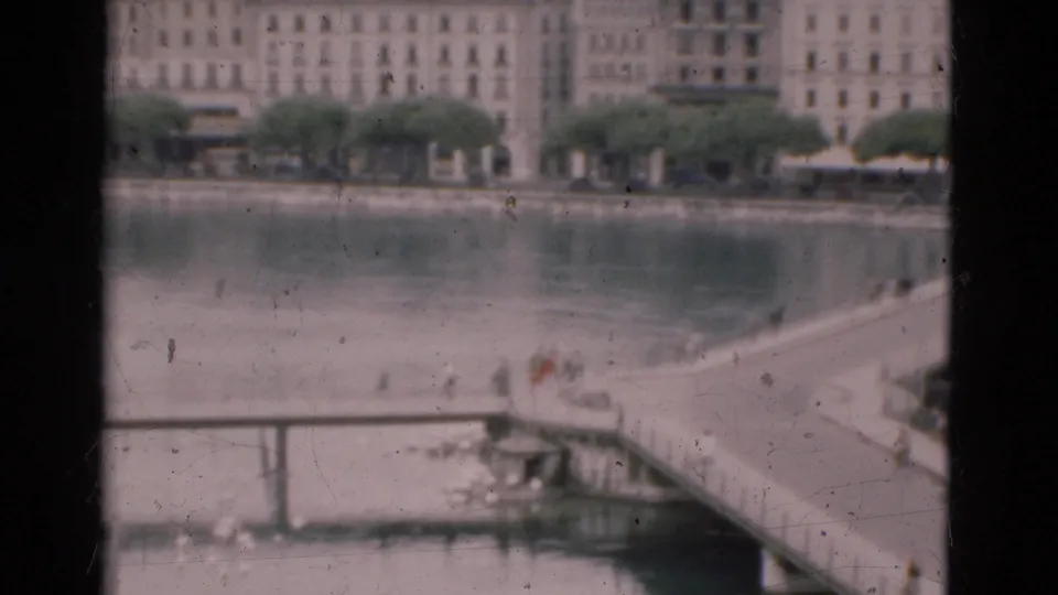 Soft 1975 Portugal home-movie frame showing riverfront buildings, water, and small pedestrians on a foreground bridge or pier.