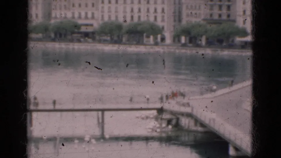 1975 Portugal home-movie frame looking through window bars toward a riverside promenade, bridge, distant walkers, trees, and far-bank buildings.