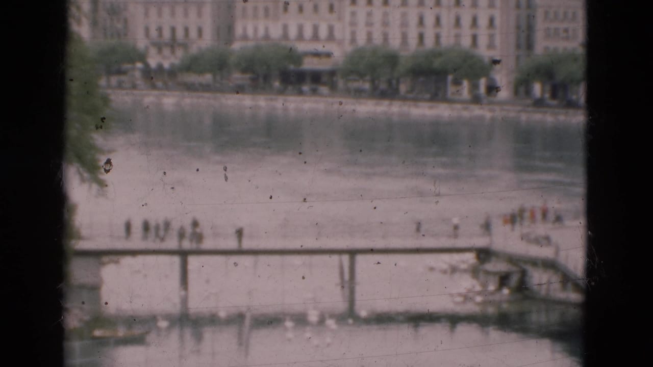 Window-framed river scene with a pedestrian bridge, small groups of people, railings, trees, and apartment buildings.