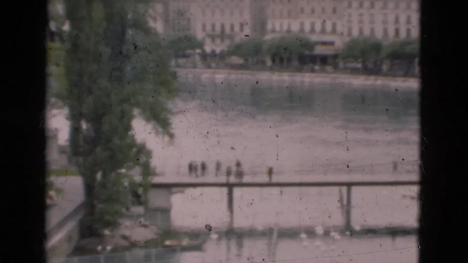 1975 Portugal 8mm frame with dark window framing, a riverside walkway, distant people, bridge structure, trees, and soft waterfront buildings.