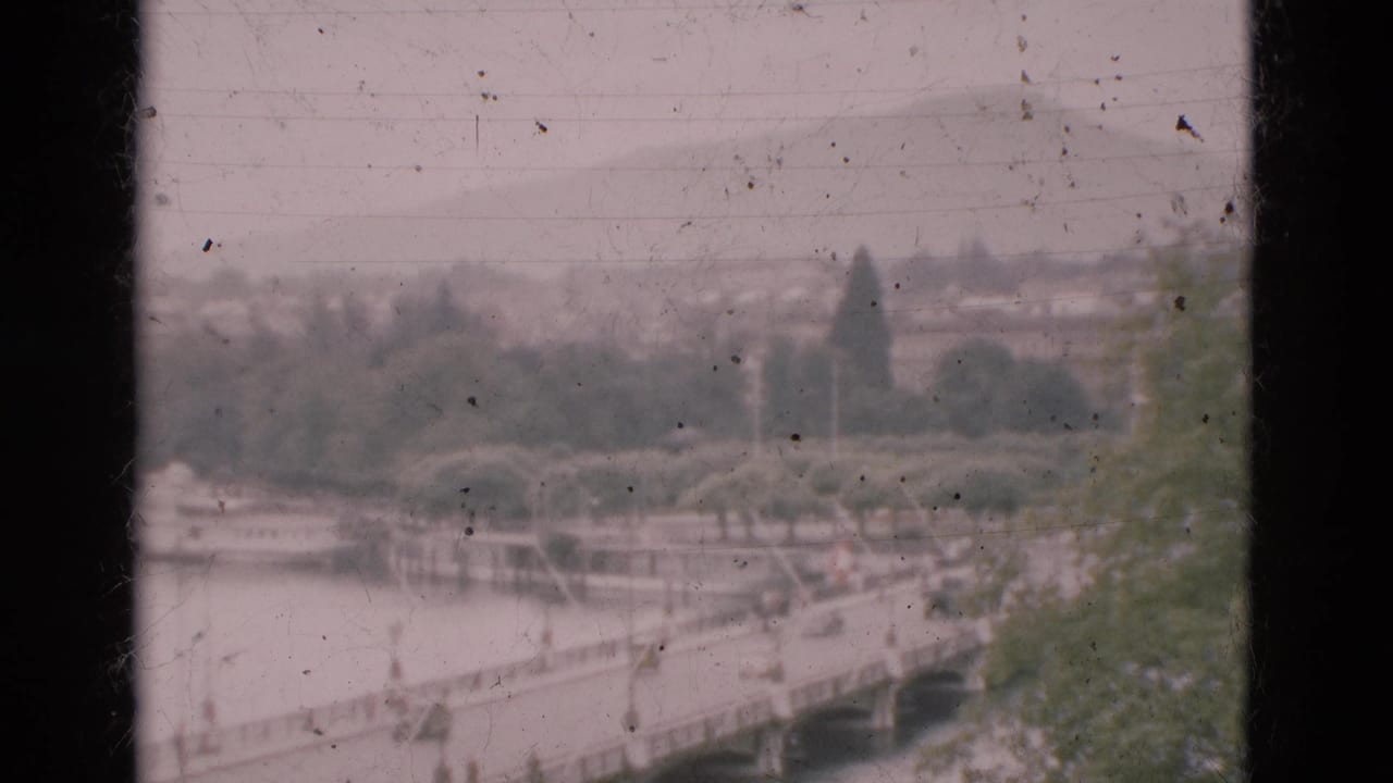 Bridge crossing a river with small cars and distant pedestrians, trees, and hazy hillside beyond.
