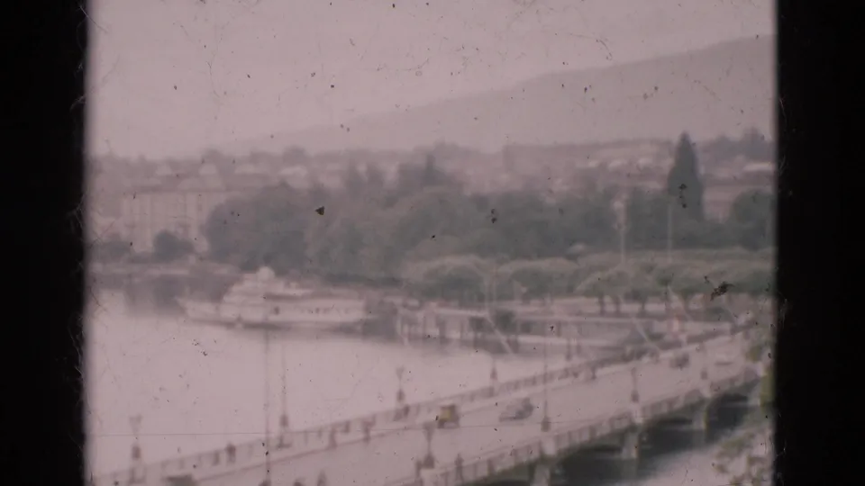 1975 Portugal home-movie frame of cars on a bridge over water, with a tree-lined bank and hazy hillside behind it.