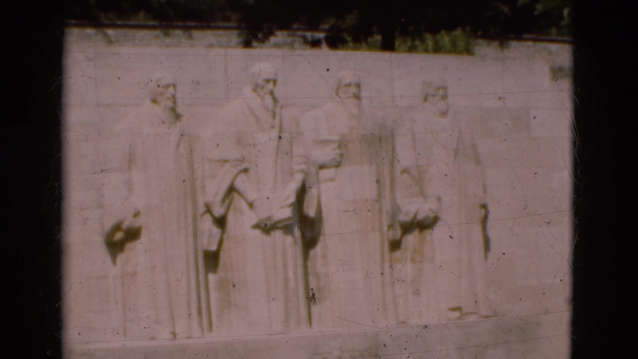 Stone relief of four standing figures on a pale wall beneath dark foliage.