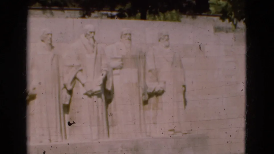 Final 8mm frame of the Portugal clip showing four solemn carved male figures in relief, foliage above, and worn film texture.