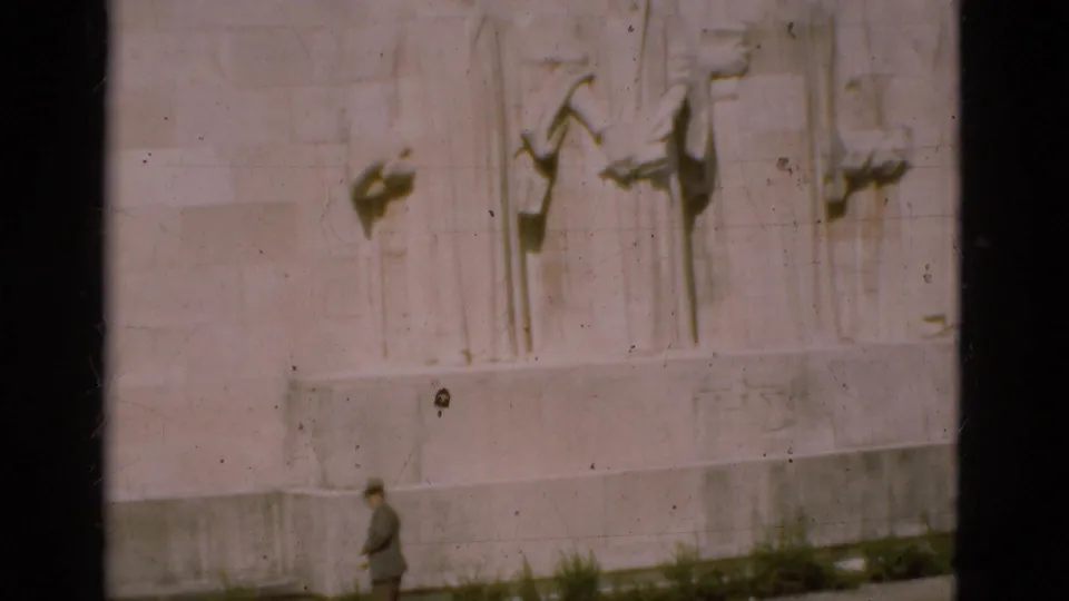1975 Portugal home-movie frame showing a man walking past the base of a pale stone bas-relief monument.