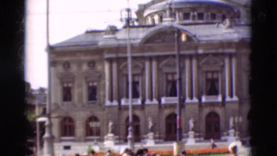 1975 Portugal 8mm frame showing the classical building across a road, with lamps, a flower bed, and pedestrians near the foreground.